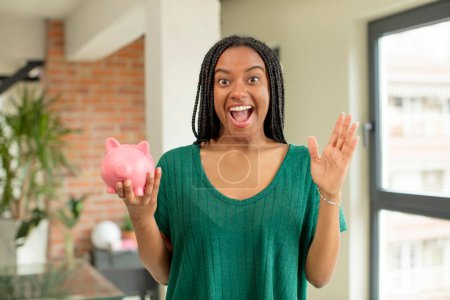 Photo for Black afro woman feeling happy and astonished at something unbelievable. piggy bank concept - Royalty Free Image