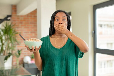 Téléchargez les photos : Afro femme noire couvrant la bouche avec une main et une expression choquée ou surprise. concept bol petit déjeuner - en image libre de droit