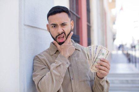 Foto de Hombre adulto joven con la boca y los ojos bien abiertos y la mano en la barbilla. concepto de billetes en dólares - Imagen libre de derechos