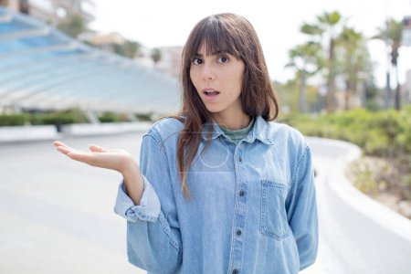 Photo for Pretty young woman looking surprised and shocked, with jaw dropped holding an object with an open hand on the side - Royalty Free Image