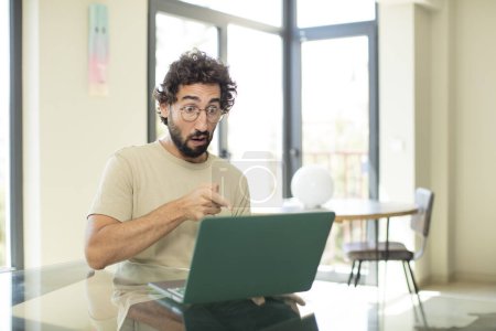 Photo for Young adult bearded man with a laptop feeling shocked and surprised, pointing and looking upwards in awe with amazed, open-mouthed look - Royalty Free Image