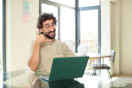 Foto de Joven hombre barbudo adulto con una computadora portátil sonriendo alegremente y apuntando a la cámara mientras hace una llamada más tarde gesto, hablando por teléfono - Imagen libre de derechos