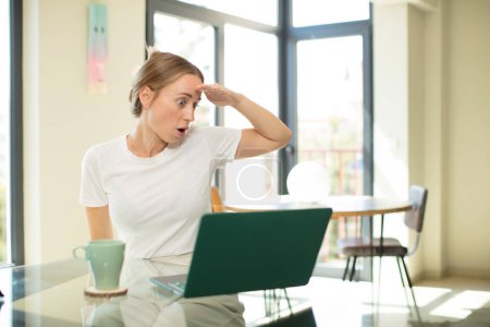 Photo for Caucasian pretty woman with a laptop looking bewildered and astonished, with hand over forehead looking far away, watching or searching - Royalty Free Image