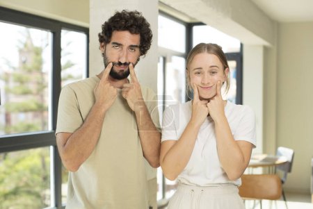 Photo for Young adult couple smiling confidently pointing to own fake smile - Royalty Free Image