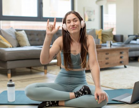 Téléchargez les photos : Jeune femme adulte pratiquant le yoga se sentant heureuse, amusante, confiante, positive et rebelle, faisant signe de rock ou de métal lourd avec la main - en image libre de droit