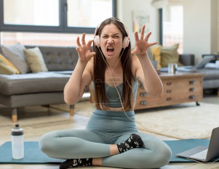 Foto de Mujer adulta joven practicando yoga gritando de pánico o rabia, conmocionada, aterrorizada o furiosa, con las manos al lado de la cabeza - Imagen libre de derechos