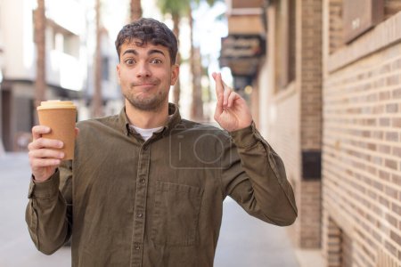 Photo for Young handsome man crossing fingers and hoping for good luck. take away coffee concept - Royalty Free Image