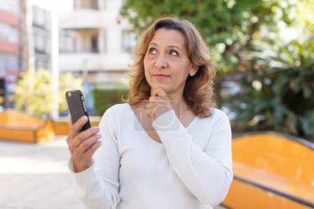 Photo for Middle age pretty woman smiling with a happy, confident expression with hand on chin. using her smartphone - Royalty Free Image