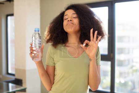 Téléchargez les photos : Jolie afro femme noire se sentant heureuse, montrant l'approbation avec le geste correct. concept de bouteille d'eau - en image libre de droit