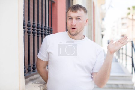 Photo for Handsome man looking surprised and shocked, with jaw dropped holding an object with an open hand on the side - Royalty Free Image