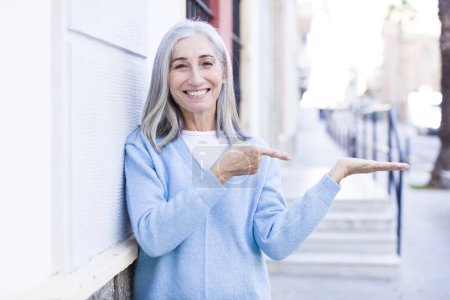 Téléchargez les photos : Senior retraité jolie femme aux cheveux blancs souriant, se sentant heureux, insouciant et satisfait, pointant vers le concept ou l'idée sur l'espace de copie sur le côté - en image libre de droit