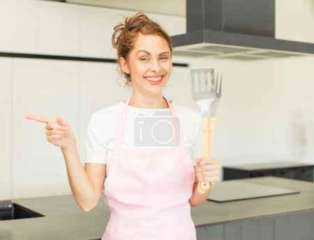 Foto de Joven bonita mujer sonriendo alegremente, sintiéndose feliz y señalando hacia un lado. concepto de chef casero - Imagen libre de derechos