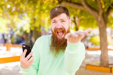Foto de Joven hombre barbudo adulto sonriendo felizmente y ofreciendo o mostrando un concepto con un teléfono inteligente - Imagen libre de derechos