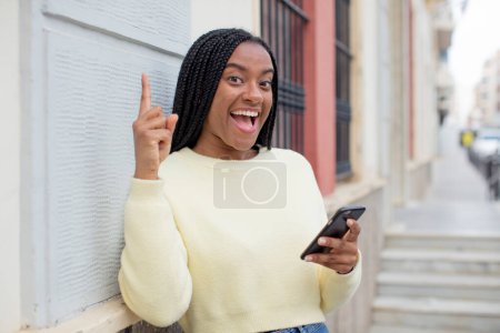 Téléchargez les photos : Femme afro noire se sentant comme un génie heureux et excité après avoir réalisé une idée. utilisant un concept de smartphone - en image libre de droit