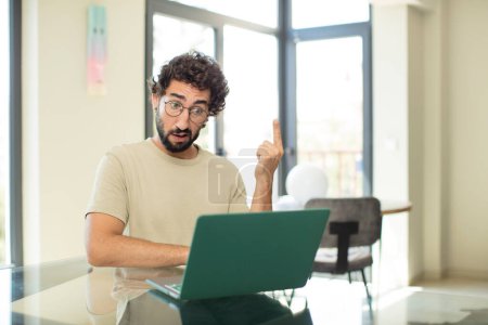 Photo for Young adult bearded man with a laptop feeling confused and puzzled, showing you are insane, crazy or out of your mind - Royalty Free Image