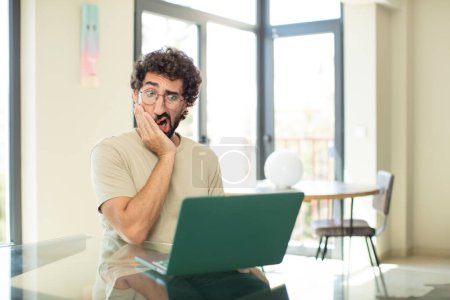 Téléchargez les photos : Jeune homme barbu adulte avec un ordinateur portable se sentant choqué et étonné tenant face à face dans l'incrédulité avec la bouche grande ouverte - en image libre de droit