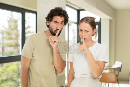 Photo for Young adult couple looking serious and cross with finger pressed to lips demanding silence or quiet, keeping a secret - Royalty Free Image