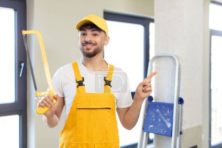 Photo for Young handsome man smiling cheerfully, feeling happy and pointing to the side. handyman with a saw - Royalty Free Image