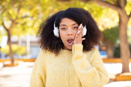 Téléchargez les photos : Jolie afro femme noire se sentant extrêmement choqué et surpris. écouter de la musique avec des écouteurs - en image libre de droit