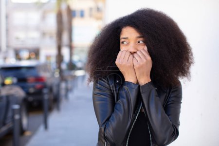 Téléchargez les photos : Afro-américaine jolie femme regardant inquiet, anxieux, stressé et effrayé, mordant les ongles et regardant vers l'espace de copie latérale - en image libre de droit