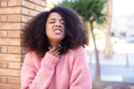 Foto de Mujer bonita afroamericana sintiéndose enferma con dolor de garganta y síntomas de gripe, tos con la boca cubierta - Imagen libre de derechos