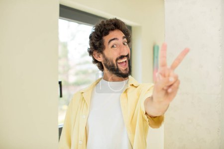 Photo for Young crazy bearded man smiling and looking happy, carefree and positive, gesturing victory or peace with one hand - Royalty Free Image
