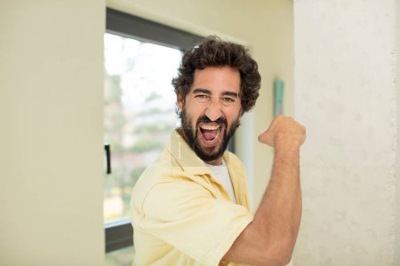 Téléchargez les photos : Jeune homme barbu fou se sentant heureux, satisfait et puissant, flexion ajustement et biceps musculaires, regardant fort après la salle de gym - en image libre de droit