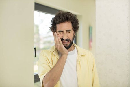 Photo for Young crazy bearded man holding cheek and suffering painful toothache, feeling ill, miserable and unhappy, looking for a dentist - Royalty Free Image