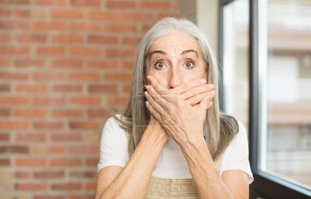 Téléchargez les photos : Senior jolie femme couvrant la bouche avec des mains avec une expression choquée et surprise, gardant un secret ou disant oups - en image libre de droit