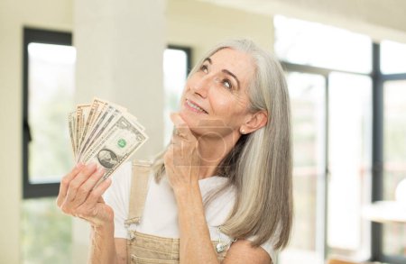 Foto de Mujer bastante mayor sonriendo con una expresión feliz y segura con la mano en la barbilla. con billetes en dólares - Imagen libre de derechos