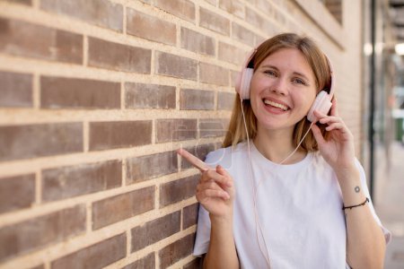 Foto de Joven bonita mujer sonriendo alegremente, sintiéndose feliz y señalando hacia un lado. auriculares y concepto de música - Imagen libre de derechos