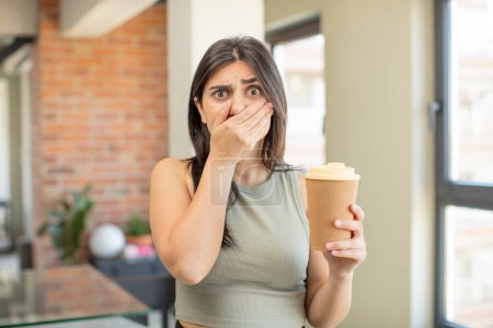 Foto de Mujer joven cubriendo la boca con una mano y expresión sorprendida o sorprendida. llevar el café - Imagen libre de derechos