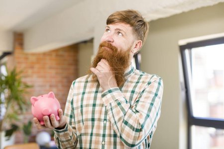 Photo for Red hair man smiling with a happy, confident expression with hand on chin with a piggy bank - Royalty Free Image