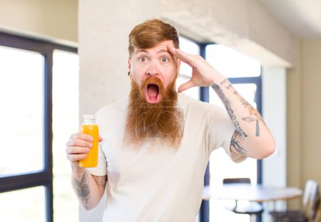 Téléchargez les photos : Homme aux cheveux roux regardant heureux, étonné et surpris avec une bouteille de jus d'orange - en image libre de droit