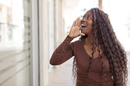 Photo for Afro pretty black woman profile view, looking happy and excited, shouting and calling to copy space on the side - Royalty Free Image