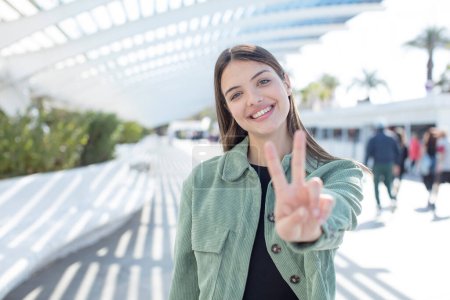 Photo for Young pretty woman smiling and looking happy, carefree and positive, gesturing victory or peace with one hand - Royalty Free Image