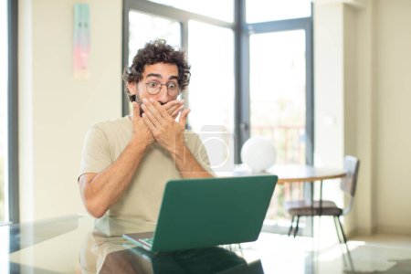 Photo for Young adult bearded man with a laptop covering mouth with hands with a shocked, surprised expression, keeping a secret or saying oops - Royalty Free Image