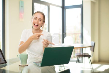 Caucasian pretty woman with a laptop smiling cheerfully and pointing to copy space on palm on the side, showing or advertising an object