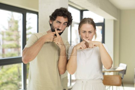 Téléchargez les photos : Jeune couple adulte regardant grave et mécontent avec les deux doigts croisés à l'avant dans le rejet, demandant le silence - en image libre de droit