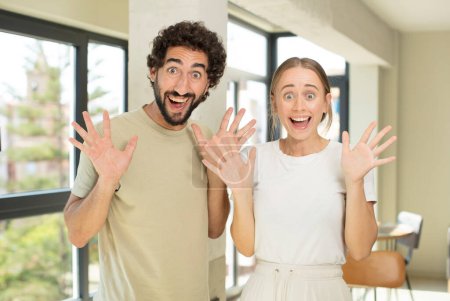 Téléchargez les photos : Jeune couple adulte regardant heureux et excité, choqué par une surprise inattendue avec les deux mains ouvertes à côté du visage - en image libre de droit