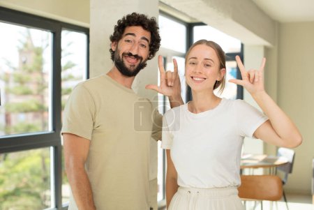 Foto de Joven pareja adulta sintiéndose feliz, divertido, seguro, positivo y rebelde, haciendo rock o heavy metal signo con la mano - Imagen libre de derechos