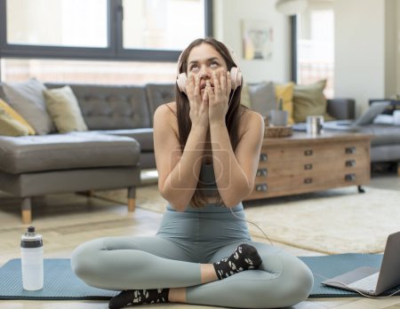 Téléchargez les photos : Jeune femme adulte pratiquant le yoga se sentant choqué et effrayé, l'air ennuyé avec les mains sur les joues - en image libre de droit