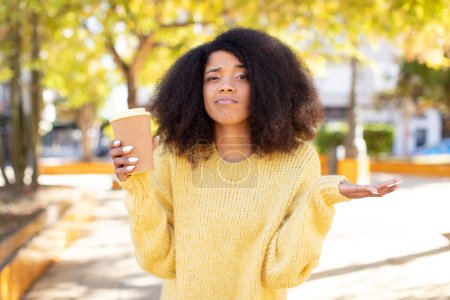 Foto de Mujer negra bastante afro encogiéndose de hombros, sintiéndose confundida e incierta. quitar el concepto de café - Imagen libre de derechos