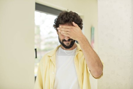 Photo for Young crazy bearded man covering eyes with one hand feeling scared or anxious, wondering or blindly waiting for a surprise - Royalty Free Image