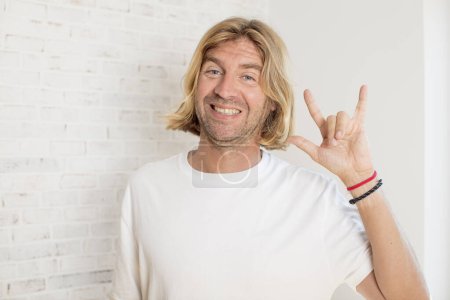 Téléchargez les photos : Jeune homme adulte blond se sentant heureux, amusant, confiant, positif et rebelle, faisant signe de rock ou de heavy metal avec la main - en image libre de droit