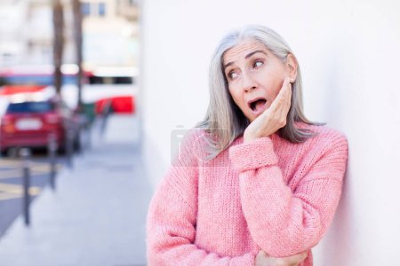 Foto de Senior jubilado bastante blanco cabello mujer sintiéndose sorprendido y asombrado sosteniendo cara a mano en la incredulidad con la boca abierta - Imagen libre de derechos