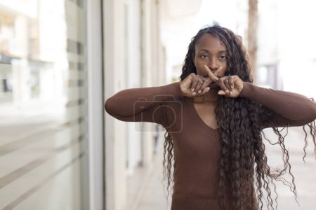 Photo for Afro pretty black woman looking serious and displeased with both fingers crossed up front in rejection, asking for silence - Royalty Free Image