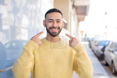 Photo for Young handsome man smiling confidently pointing to own broad smile, positive, relaxed, satisfied attitude - Royalty Free Image