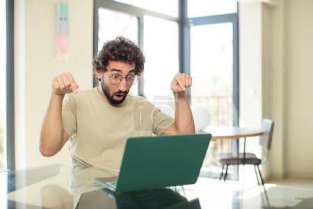 Photo for Young adult bearded man with a laptop with open mouth pointing downwards with both hands, looking shocked, amazed and surprised - Royalty Free Image