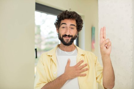Photo for Young crazy bearded man looking happy, confident and trustworthy, smiling and showing victory sign, with a positive attitude - Royalty Free Image